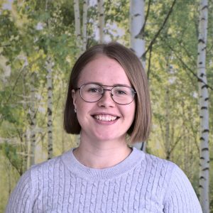Julia Lyytinen hymyilee. Taustalla seinä, jossa koivumetsäkuviointi