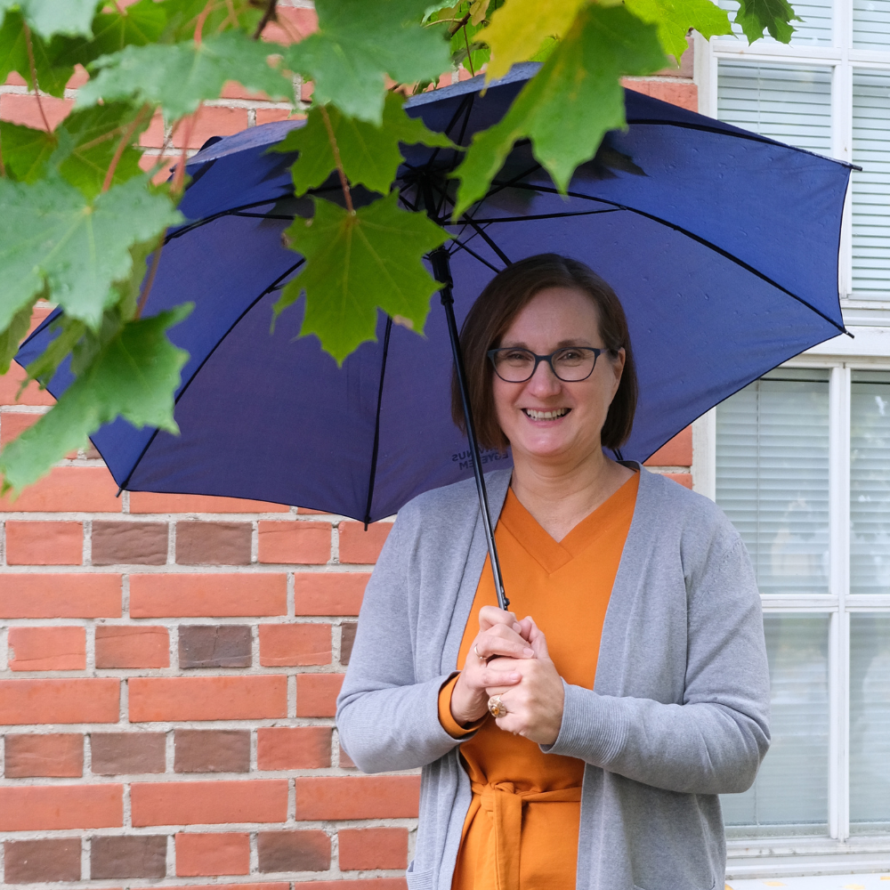 Kuvassa Marjo Juuti-Tavi TATUn toiminnanjohtaja ja järjestösuunnittelija pitelee sateenvarjoa ja hymyilee.