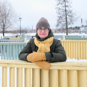 Toiminnanjohtaja/järjestösuunnittelija Marjo Juuti-Tavi lumisessa puistossa, keltainen huivi kaulassa nojaa aitaan ja hymyilee.