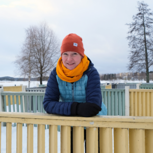Järjestösuunnittelija Tiina Männistö lumisessa puistossa, keltainen huivi kaulassa nojaa aitaan ja hymyilee.