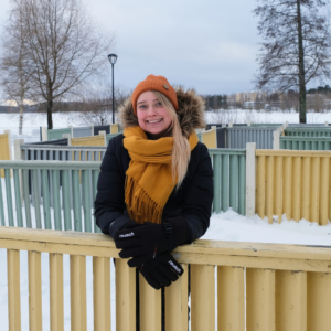 VAUHTI!-suunnittelija Tessa Ollila lumisessa puistossa, keltainen huivi kaulassa nojaa aitaan ja hymyilee.
