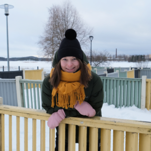 VAUHTI!-suunnittelija Katja Ihalainen lumisessa puistossa, keltainen huivi kaulassa nojaa aitaan ja hymyilee.