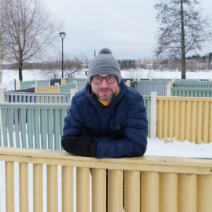 VAUHTI!-suunnittelija Petri Partanen lumisessa puistossa, keltainen huivi kaulassa nojaa aitaan ja hymyilee.
