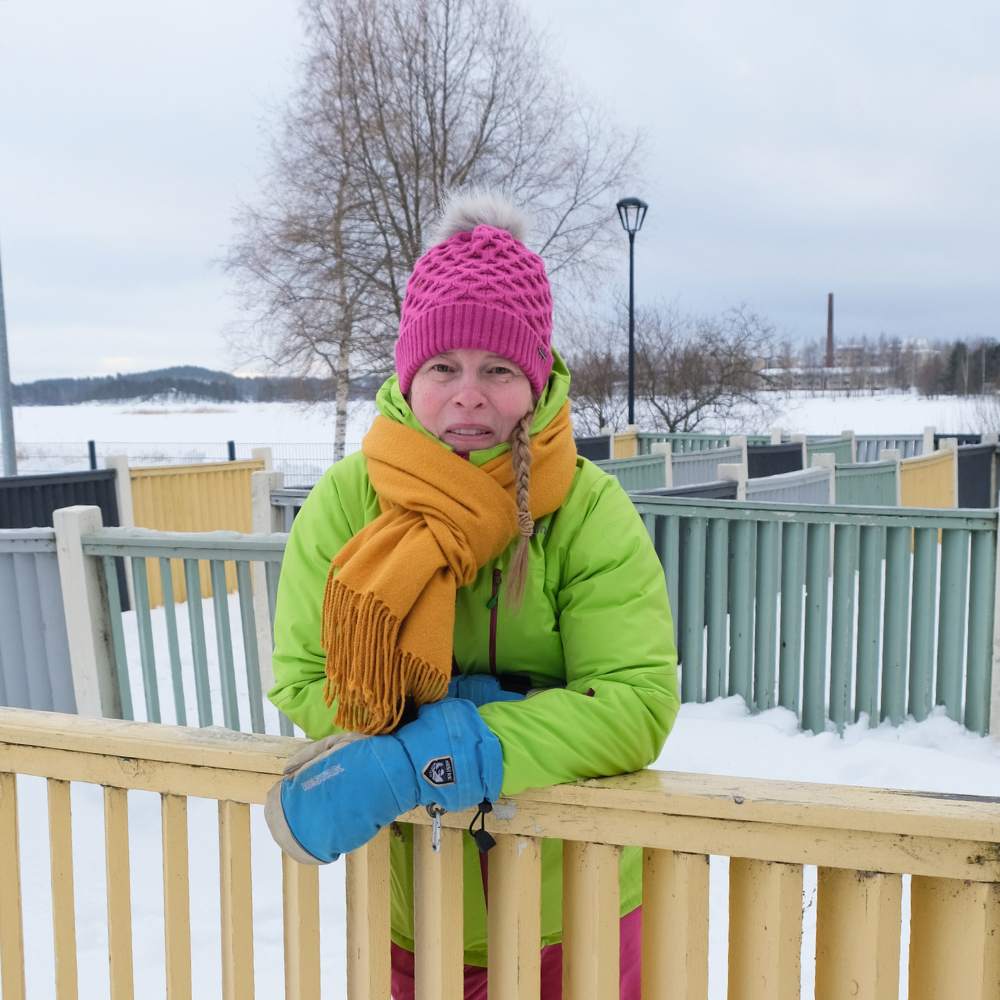 Järjestösuunnittelija Anu Hämäläinen lumisessa puistossa, keltainen huivi kaulassa nojaa aitaan ja hymyilee.