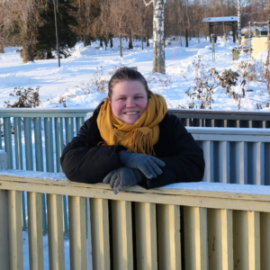 Kurssisuunnittelija Emma Lappi lumisessa puistossa, keltainen huivi kaulassa nojaa aitaan ja hymyilee.