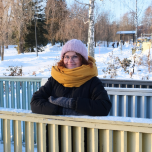 Viestintäsuunnittelija Eija Saario lumisessa puistossa, keltainen huivi kaulassa nojaa aitaan ja hymyilee.